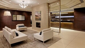 A modern lobby with two white sofas, a floor lamp, a fireplace, and a reception desk with a wooden backdrop. - Cambridge Suites Hotel (Halifax)