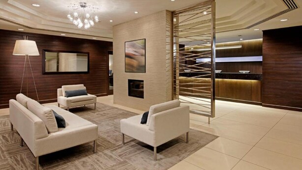 A modern lobby with two white sofas, a floor lamp, a fireplace, and a reception desk with a wooden backdrop. - Cambridge Suites Hotel (Halifax)