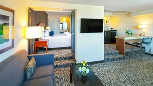 A hotel room with a bed, a sofa, a coffee table with flowers, a TV, a desk, and a kitchen area. - Cambridge Suites Hotel (Halifax)