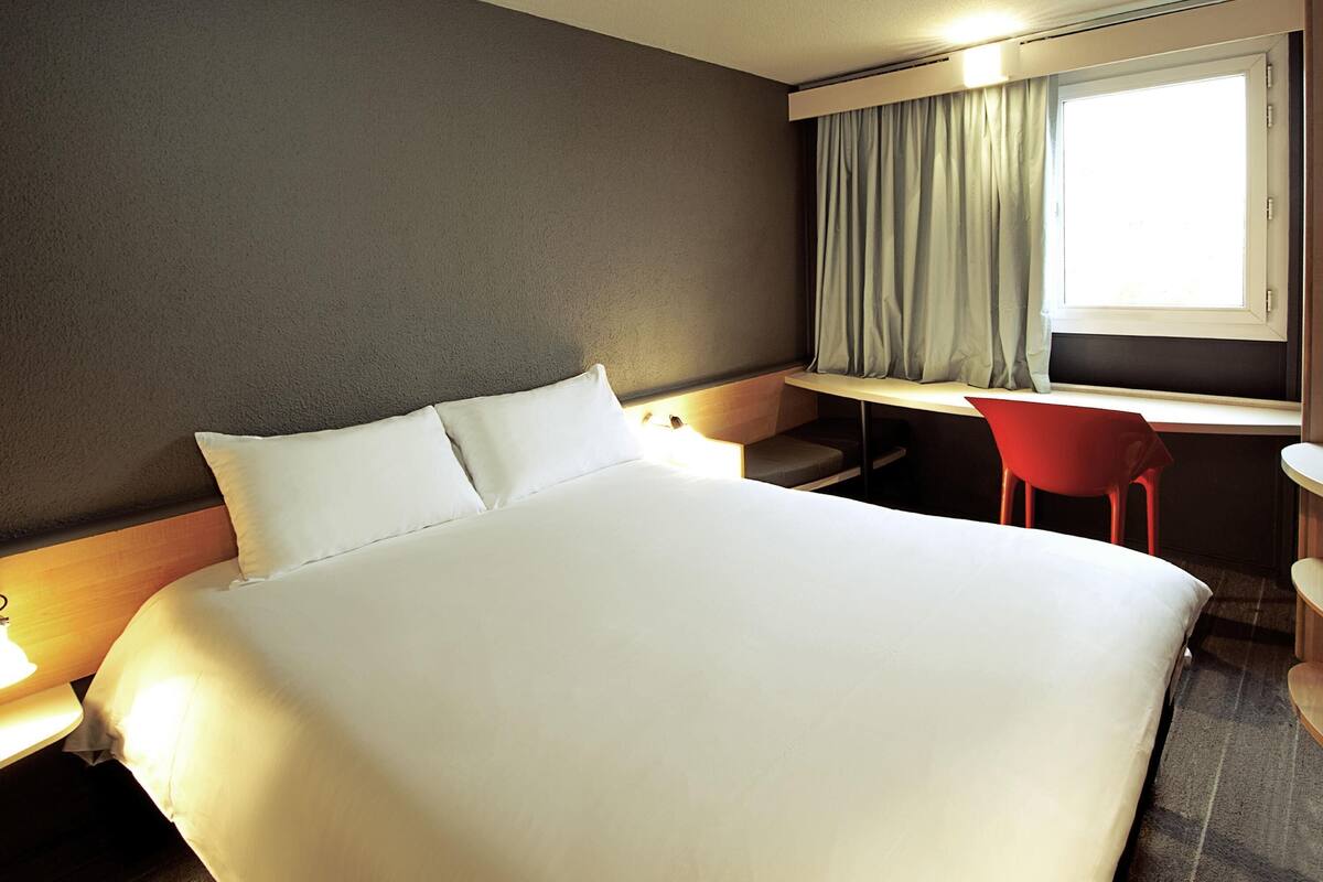 A hotel room with a large bed, a desk, a chair, and a window with curtains.