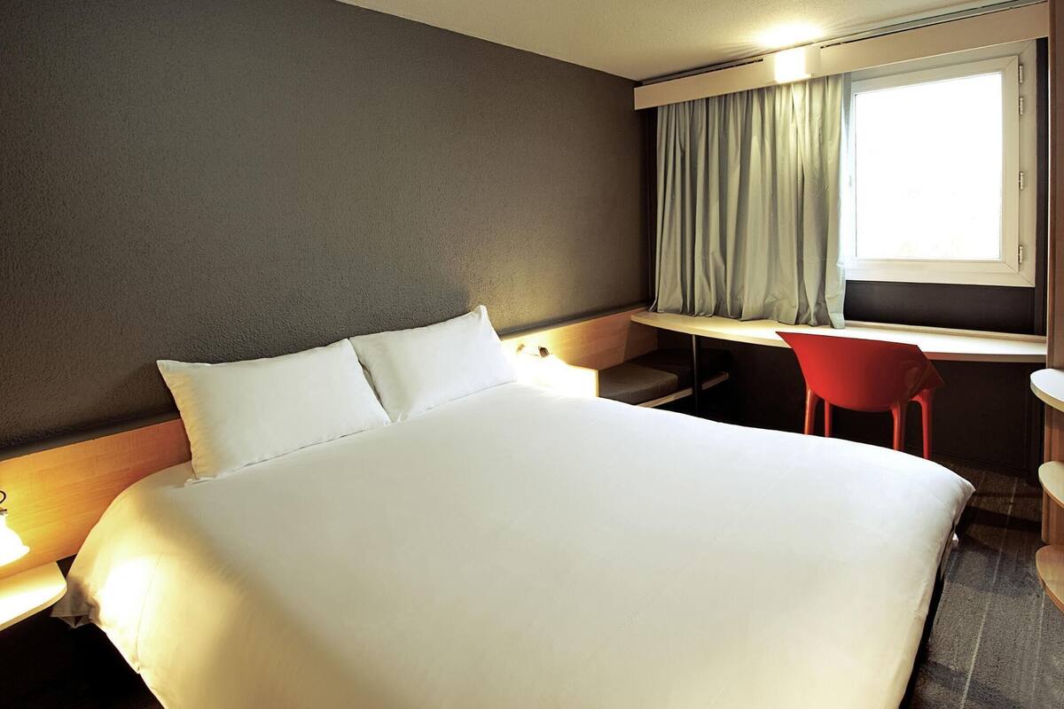 Una habitación de hotel con cama, escritorio, silla y ventana con cortinas.