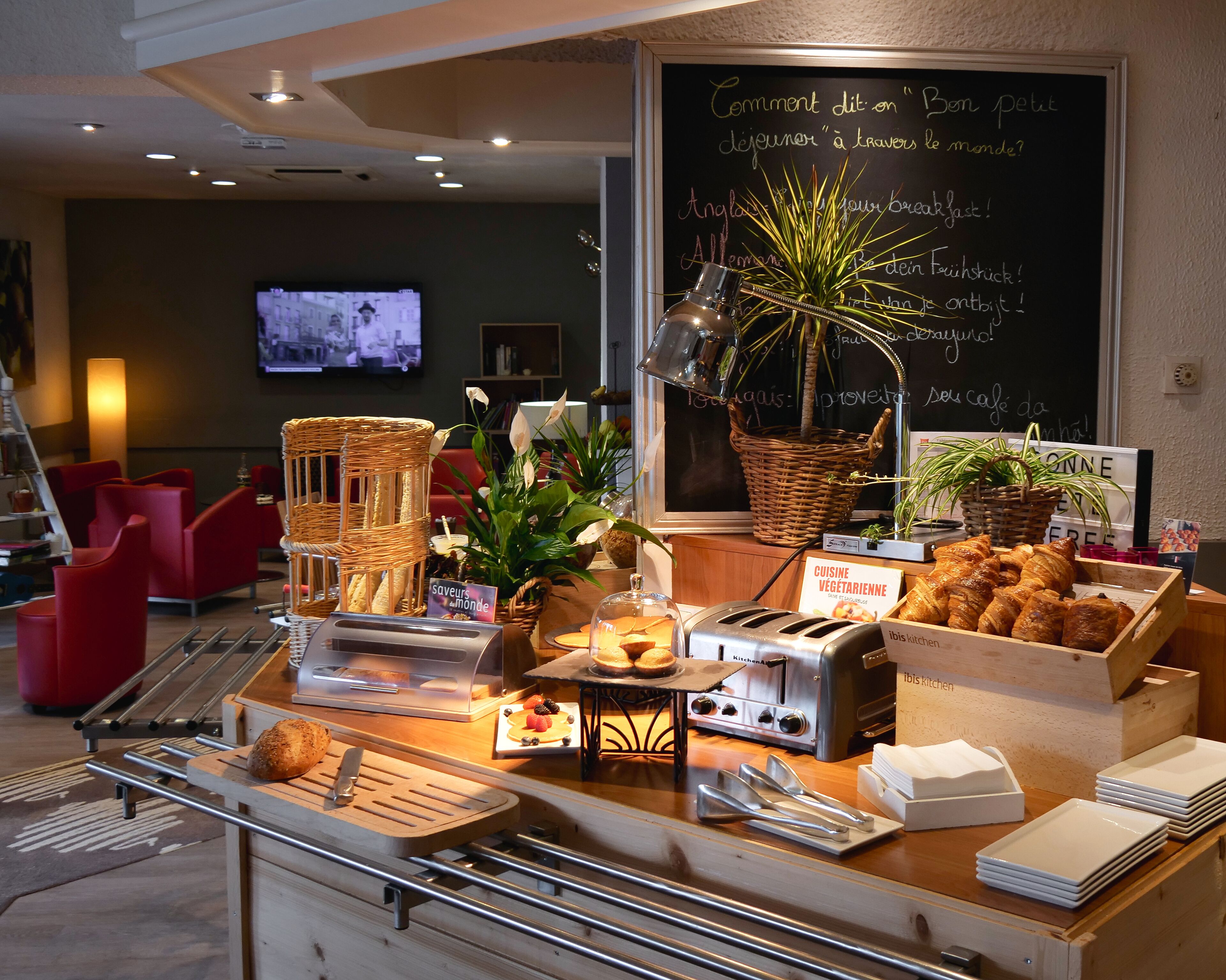 A breakfast buffet with various food items, a chalkboard with menu items, and a television in the background.