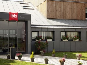 A modern hotel with a glass facade, a red ibis logo, and potted plants on the exterior. - Hotel ibis Nemours (Nemours)