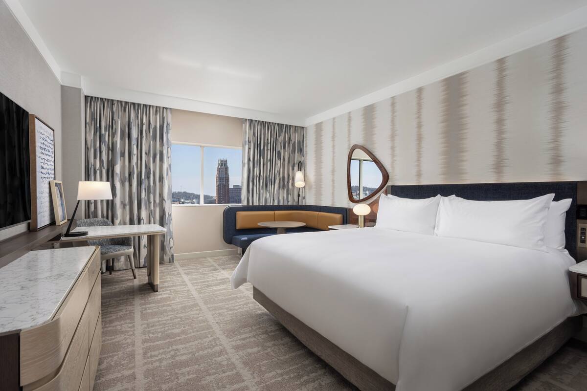 A hotel room with a large bed, a desk, a chair, a sofa, and a view of a cityscape.