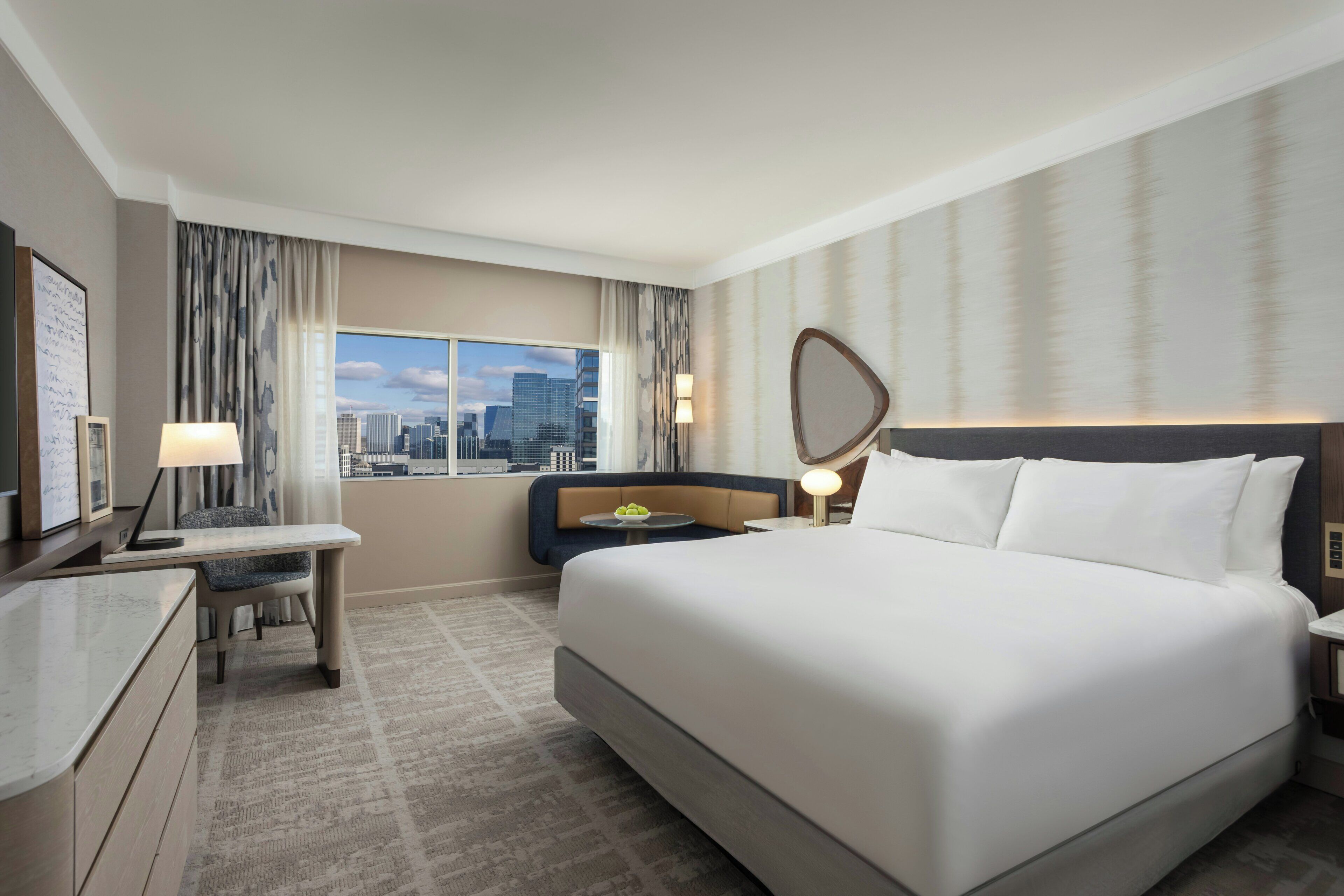 A hotel room with a large bed, a desk, a chair, a bedside table with a lamp, a view of the cityscape, and a large mirror on the wall.