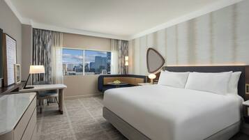 A hotel room with a large bed, a desk, a chair, a bedside table with a lamp, a view of the cityscape, and a large mirror on the wall.