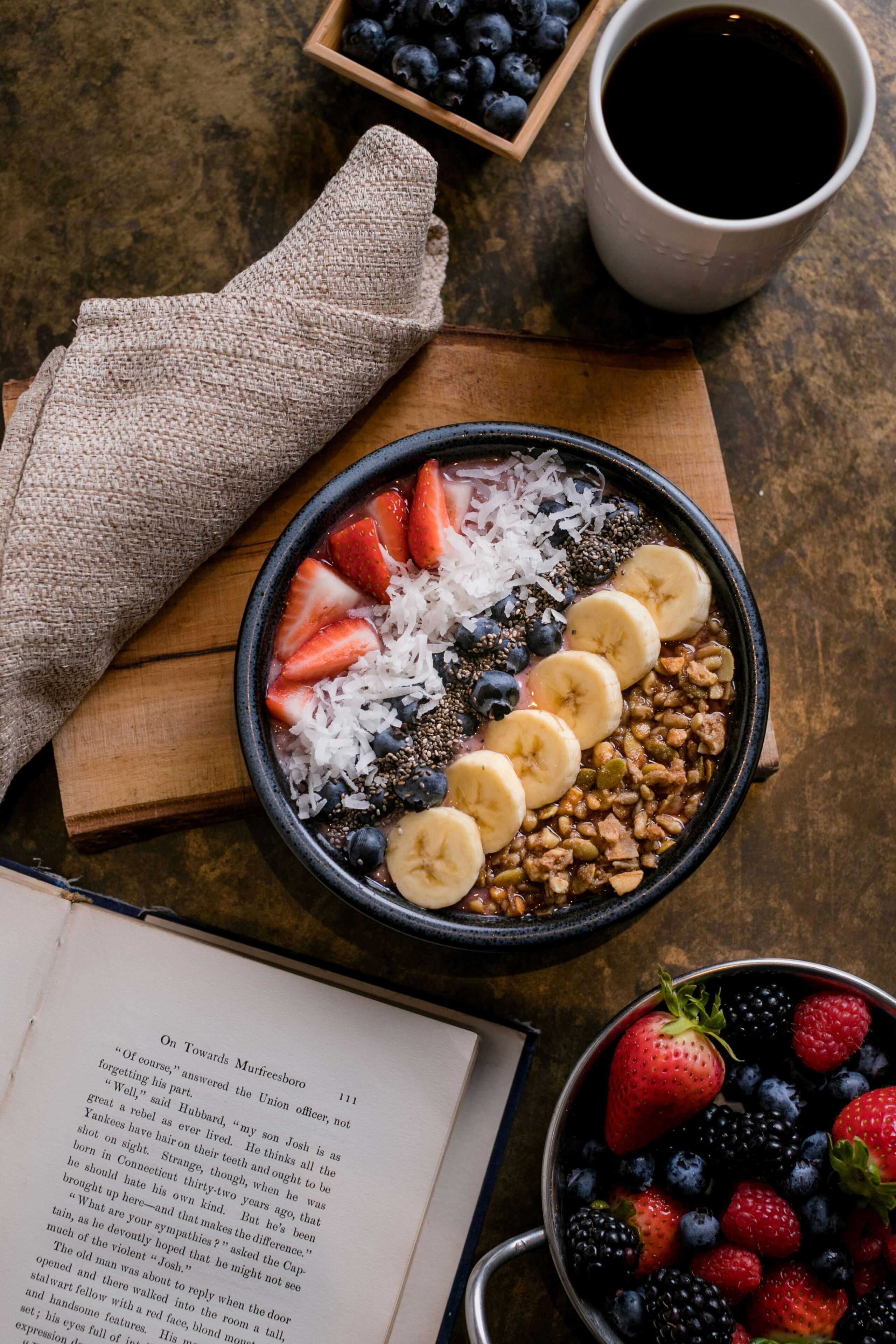 Een kom fruit en granola op een houten plank, een kop koffie, en een boek op een tafel.