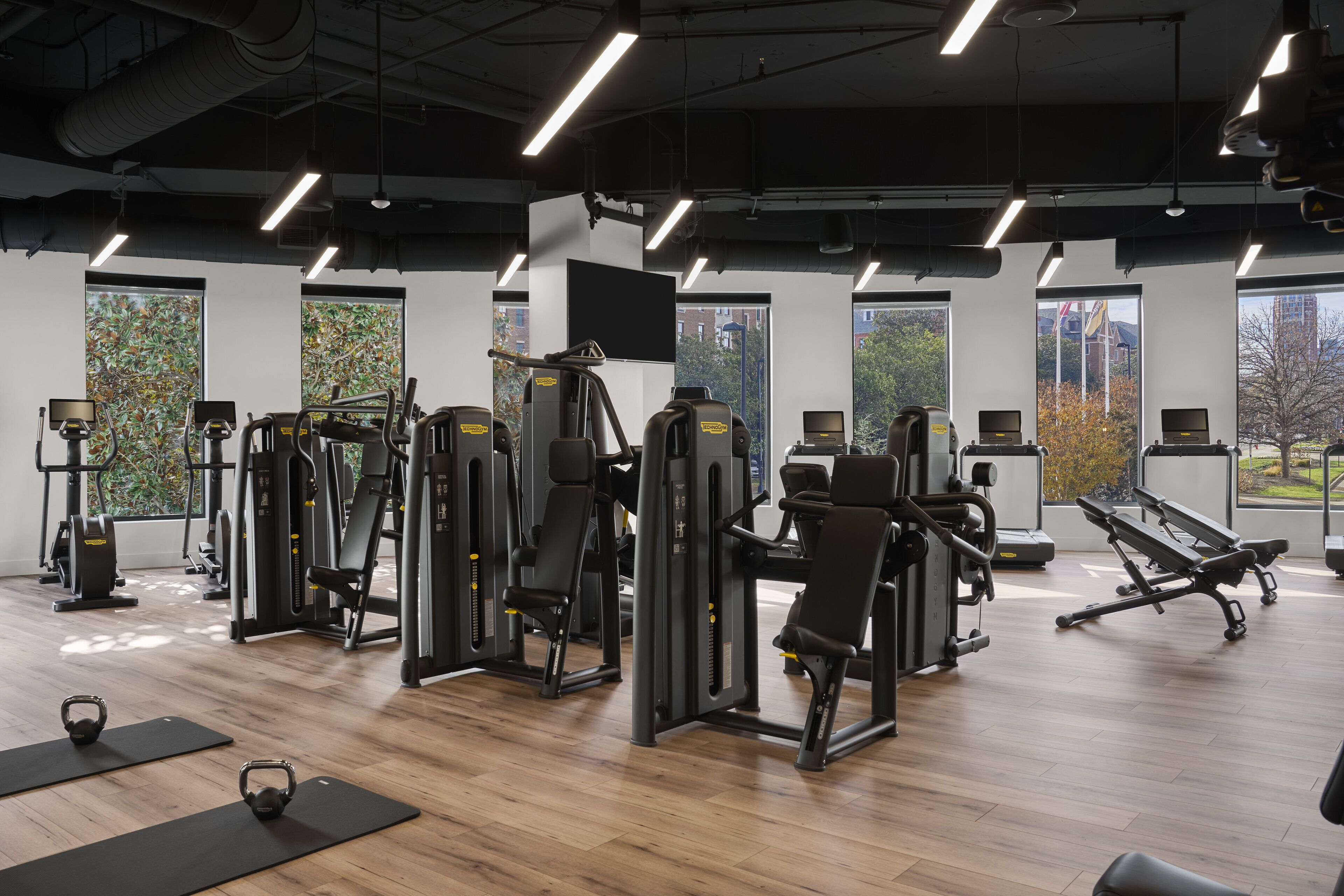 A gym with various exercise machines, wooden flooring, and large windows offering views of the outdoors.