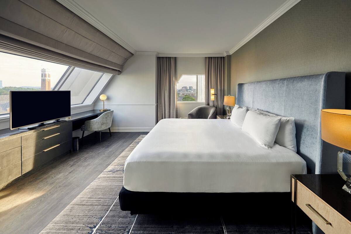 A hotel room with a large bed, a desk with a flat-screen TV, a chair, and a view of the city through the window.