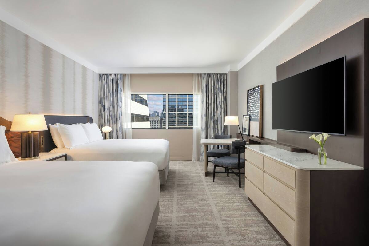 A hotel room with two beds, a desk, a TV, and a view of the city.