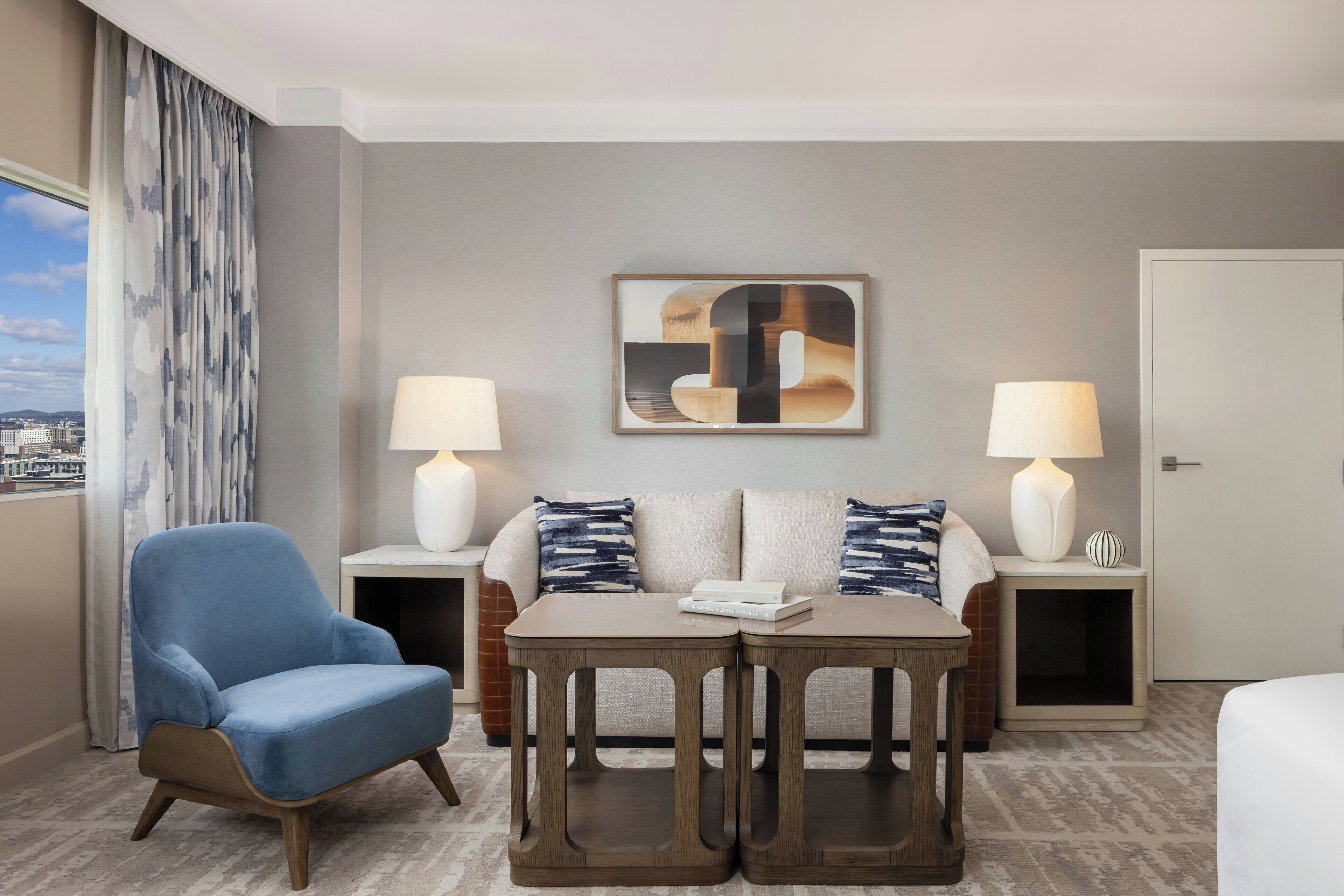 A hotel room with a blue armchair, a sofa with patterned pillows, a coffee table, two bedside lamps, and a large abstract artwork on the wall.