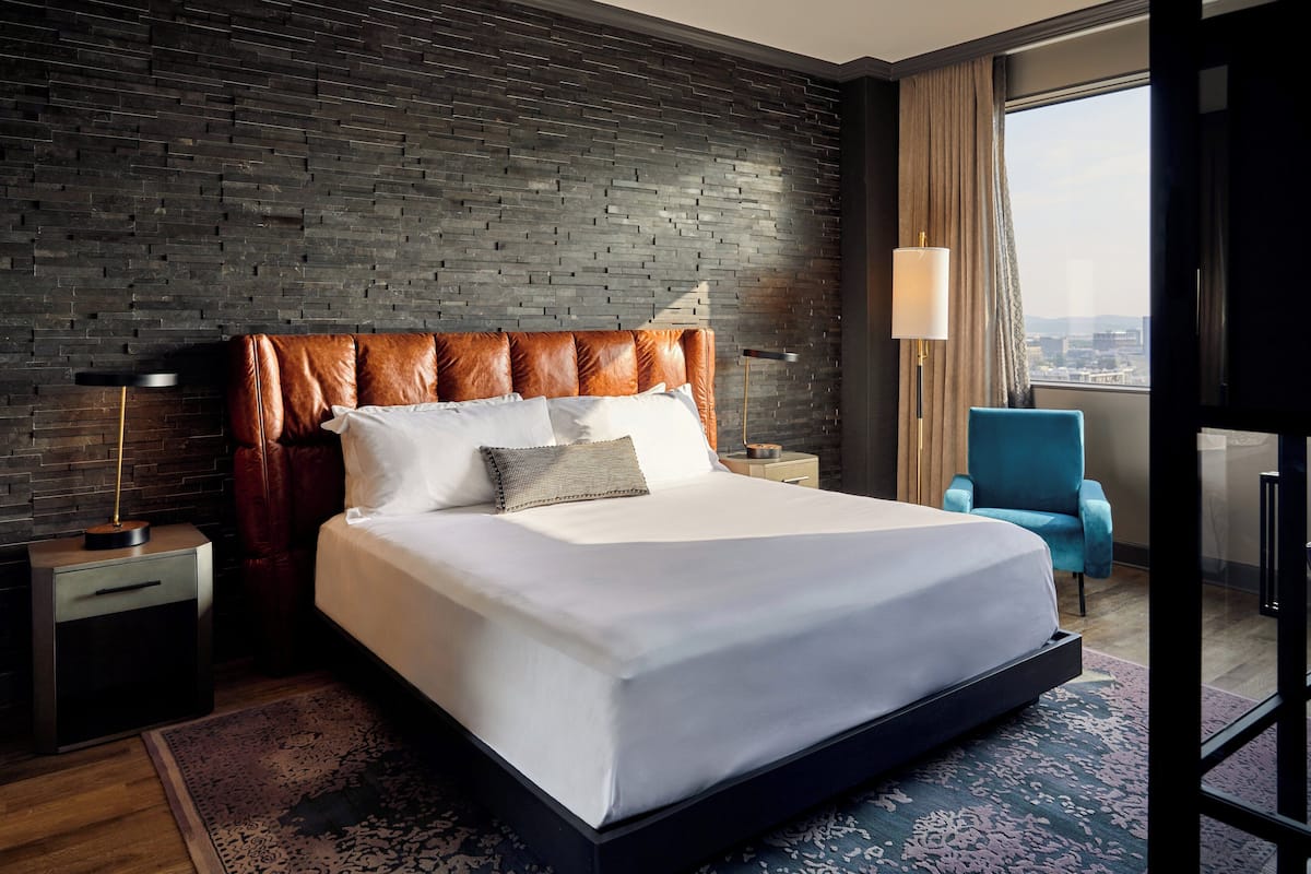 A modern hotel room with a large bed, a brick-patterned wall, a view of the city, and a blue armchair.