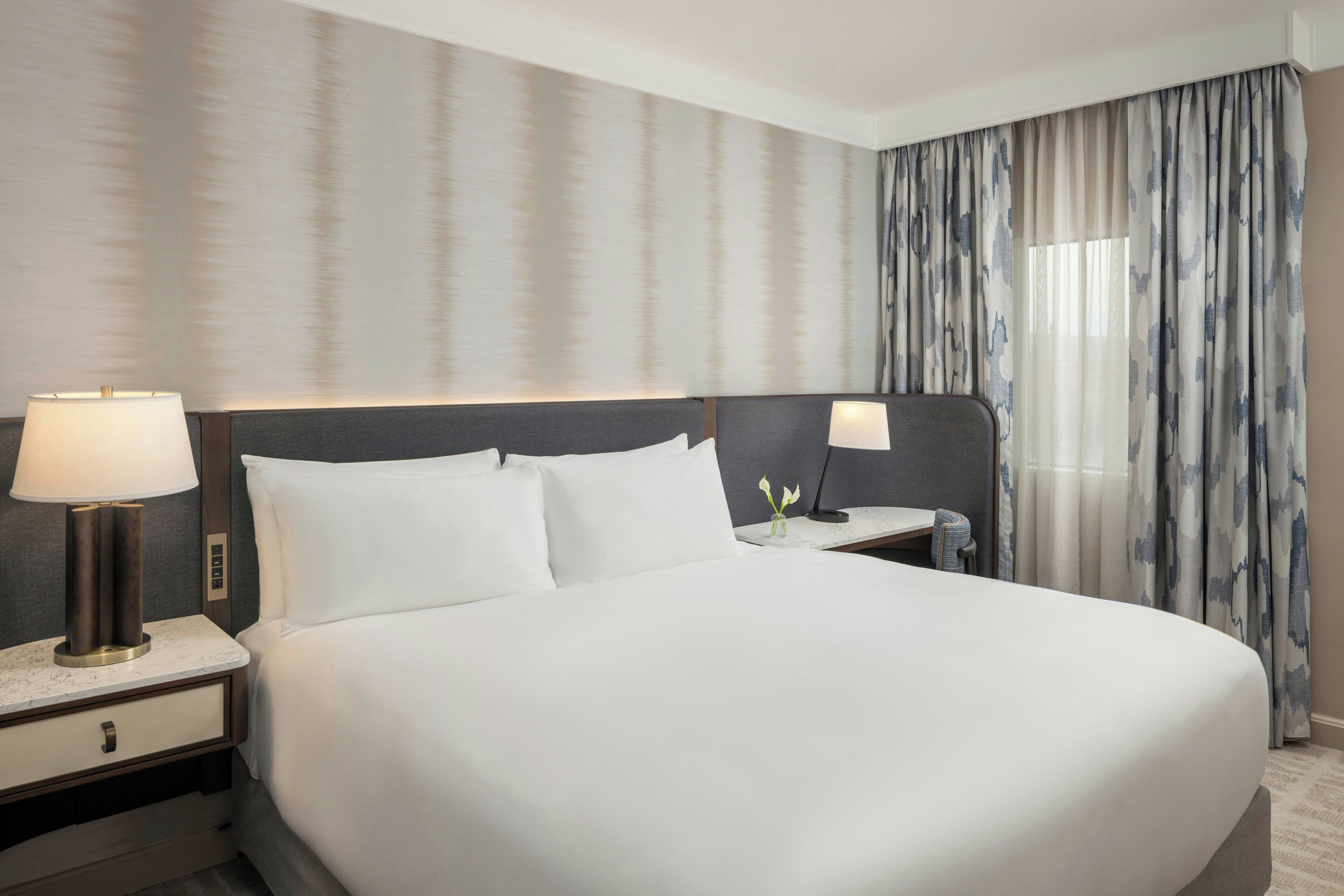 A hotel room with a large bed, two bedside tables with lamps, a nightstand, and a window with curtains.