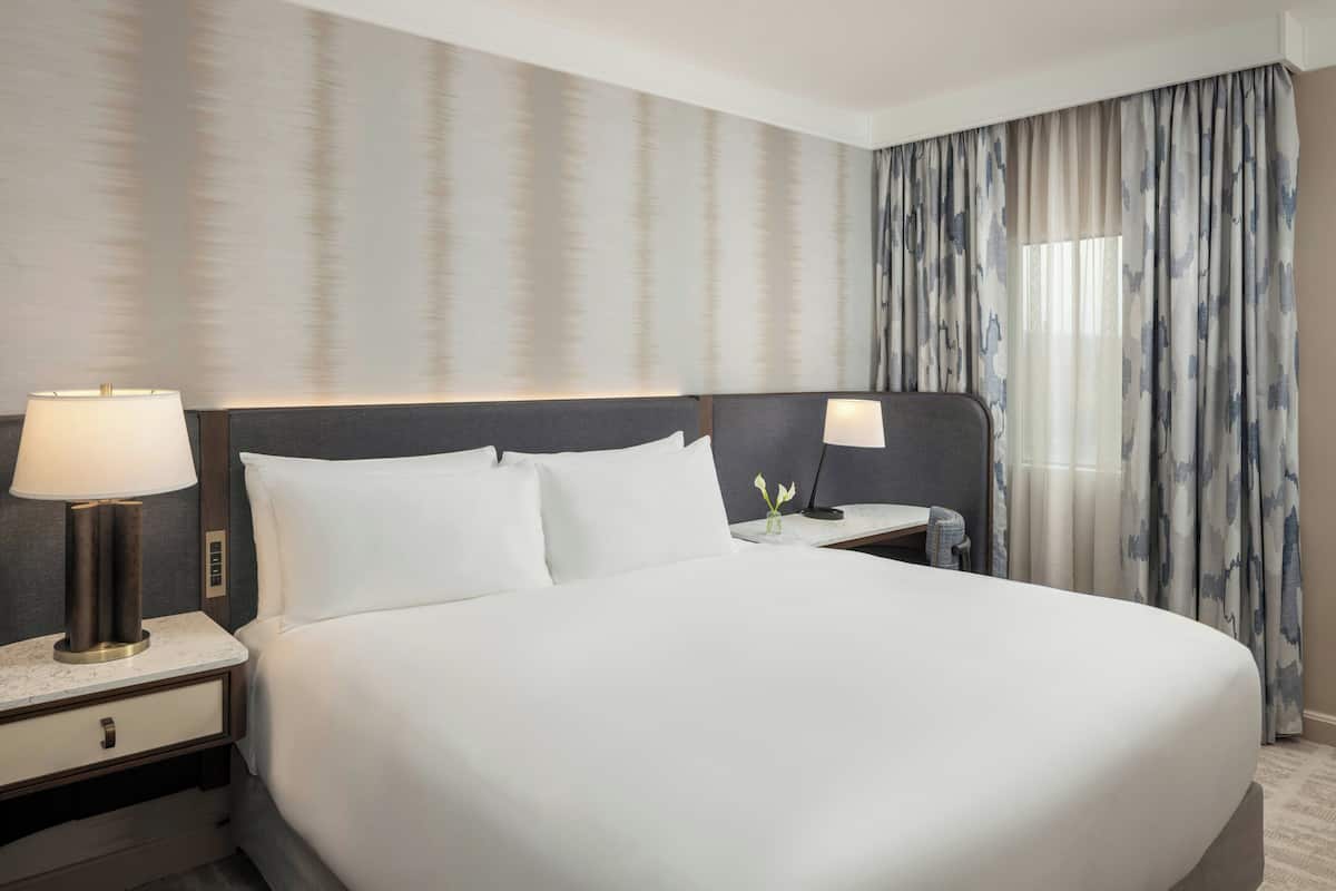 A hotel room with a large bed, two bedside tables with lamps, a nightstand, and a window with curtains.