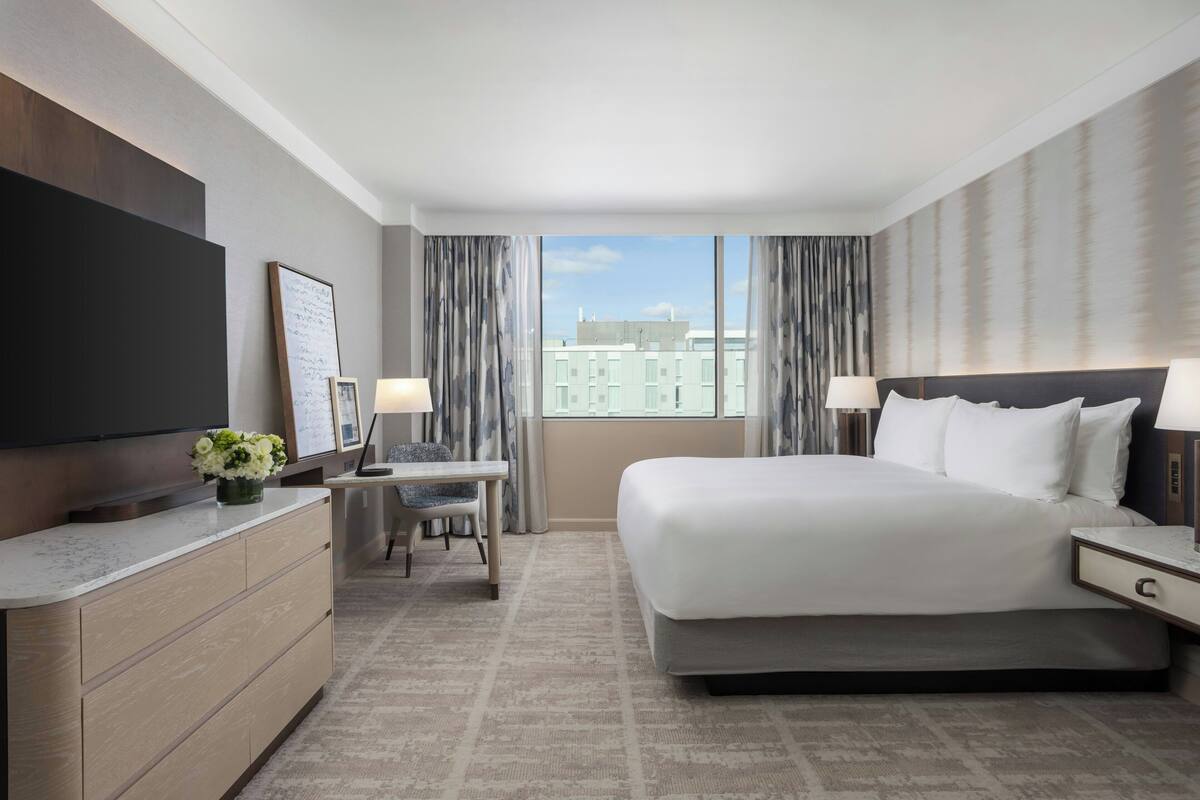 A hotel room with a large bed, a desk with a chair, a flat-screen TV, and a window with curtains.