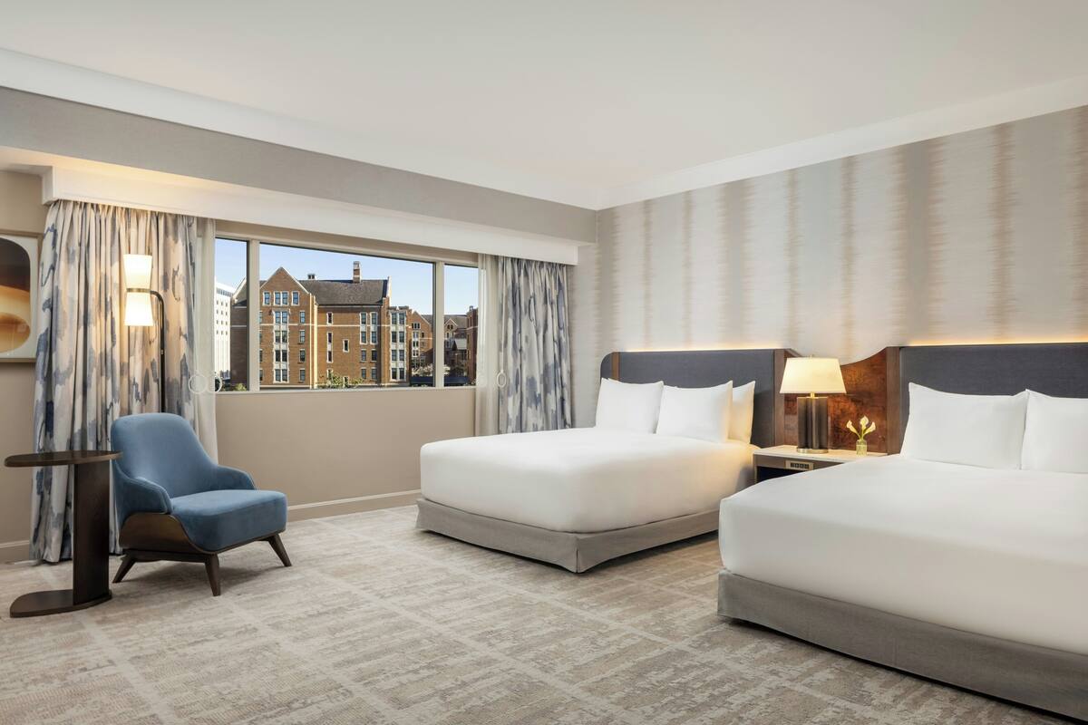 A hotel room with two beds, a chair, a nightstand, and a view of buildings.
