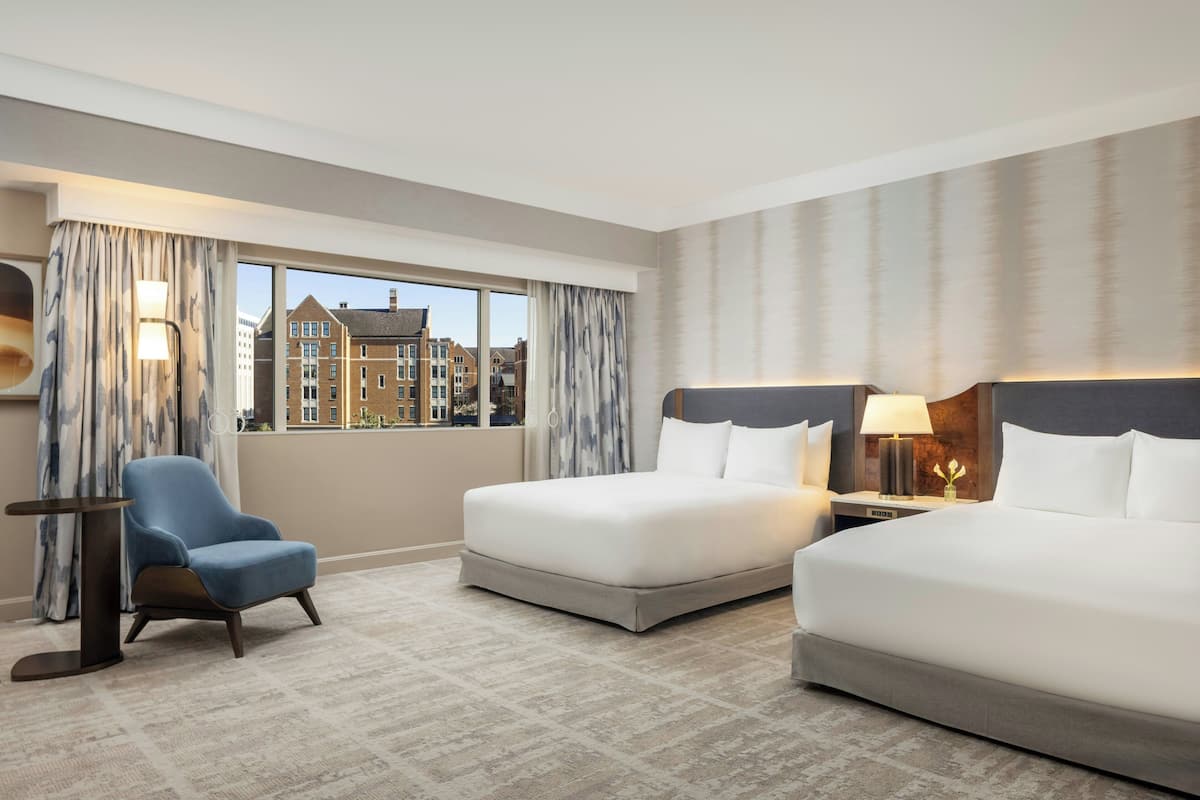 A hotel room with two beds, a chair, a nightstand, and a view of buildings.