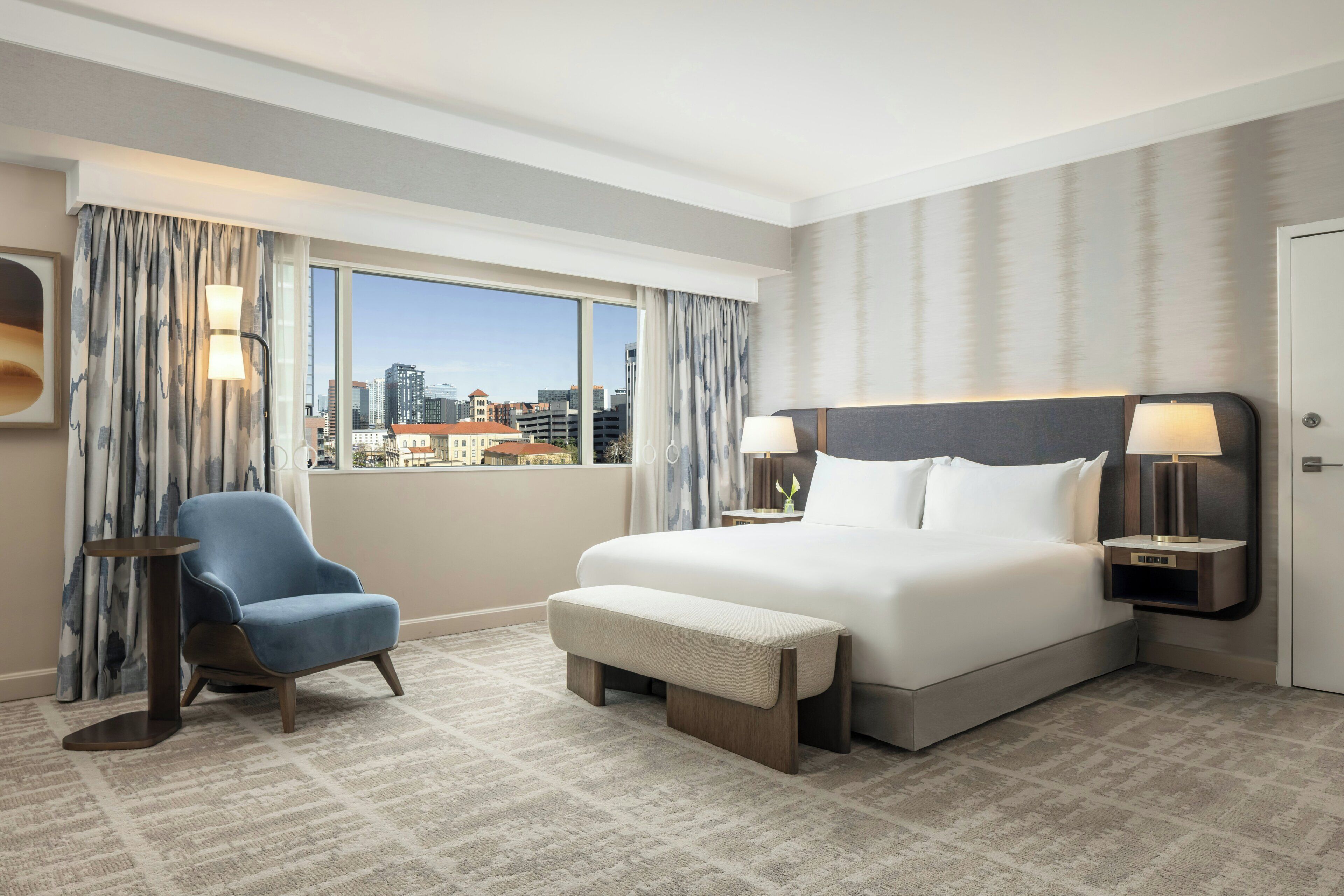 A hotel room with a large bed, a chair, a bench, two bedside tables with lamps, and a view of the city through large windows.