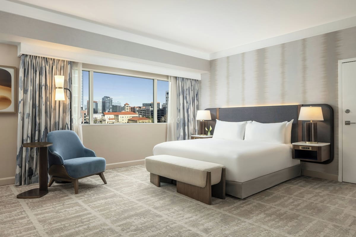 A hotel room with a large bed, a chair, a bench, two bedside tables with lamps, and a view of the city through large windows.