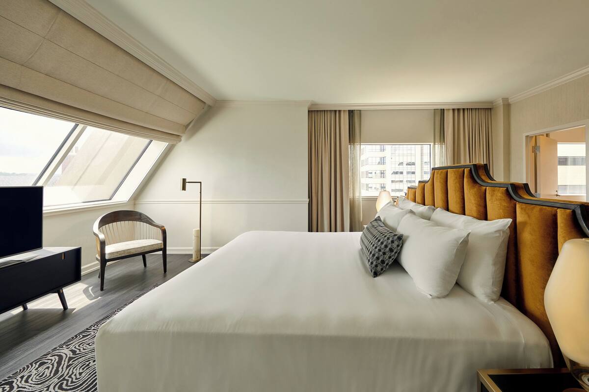 A hotel room with a large bed, a television, a chair, and a view of the city through the window.
