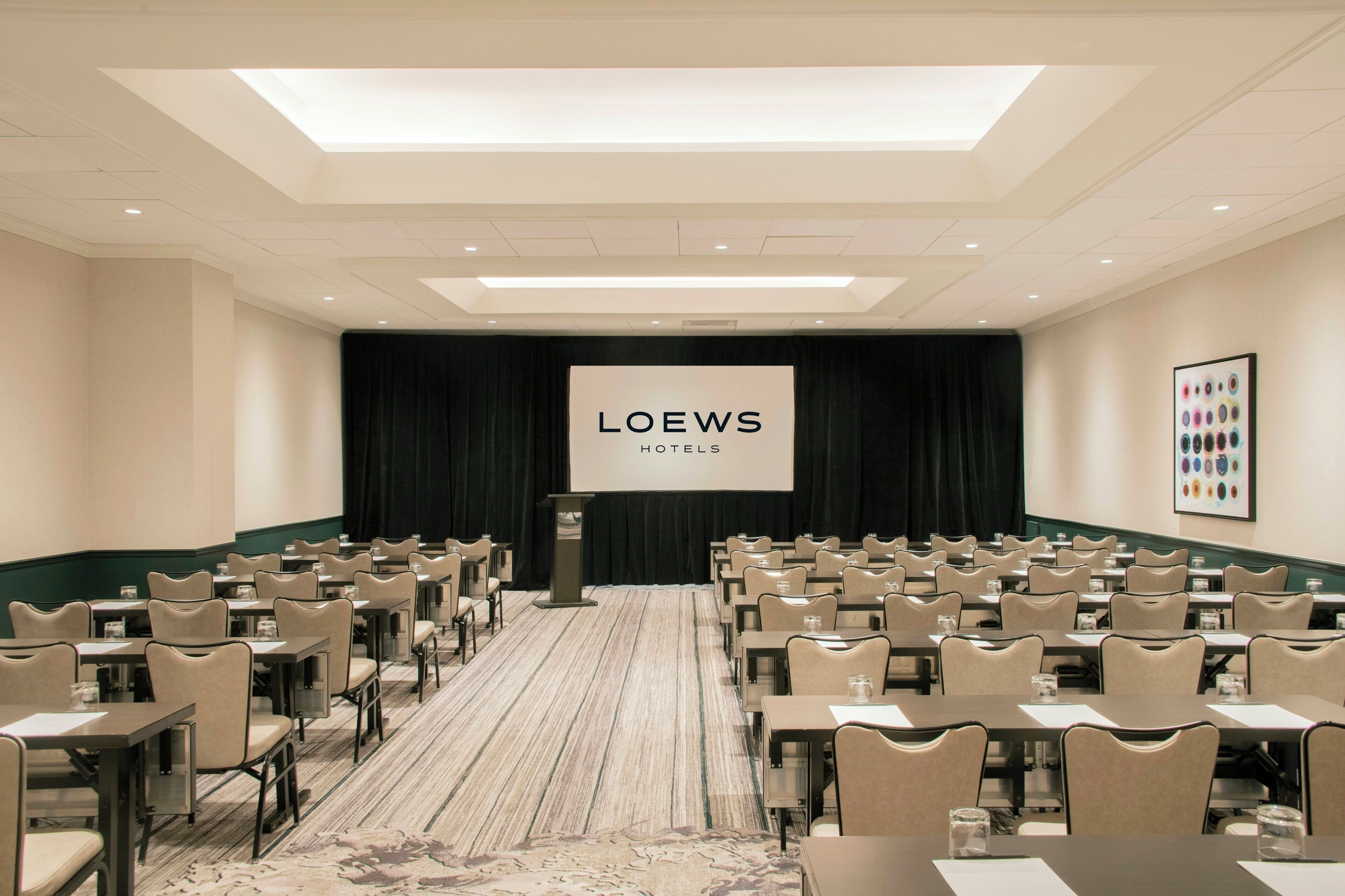 A conference room with rows of tables and chairs, a podium with a screen displaying 'LOEWS HOTELS', and a framed picture on the wall.