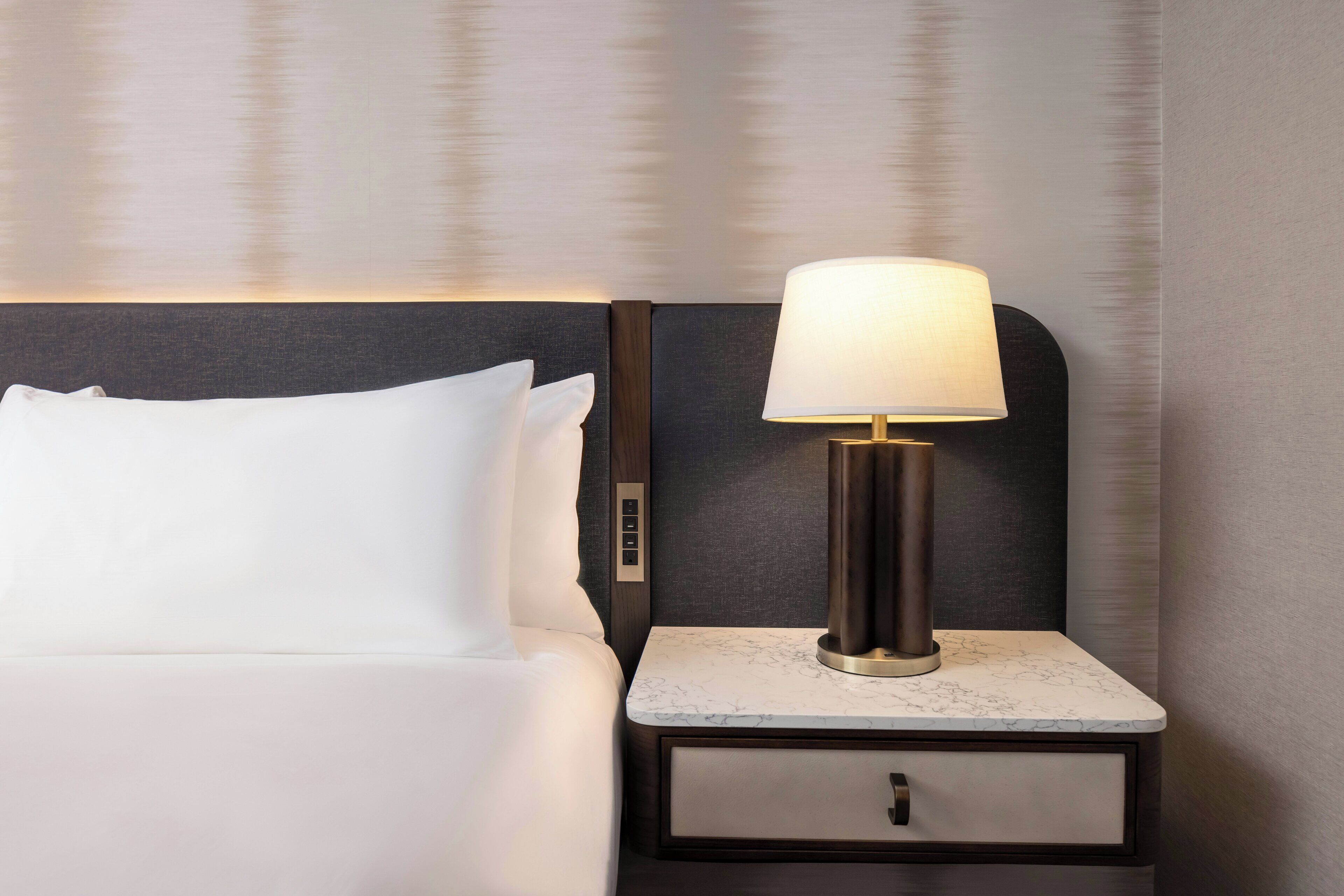 A neatly made bed with white linens, a bedside table with a lamp, and a small drawer.