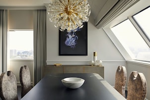 A modern dining room with a dark table, a white bowl, and unique chairs. A chandelier hangs from the ceiling, and a framed artwork is on the wall.