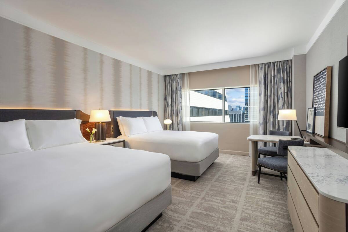 A hotel room with two beds, a desk, a chair, a TV, and a window with a city view.