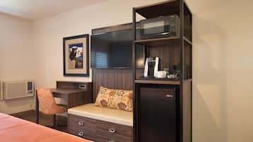A compact hotel room with a bed, a desk with a chair, a microwave, a refrigerator, and a framed picture on the wall.