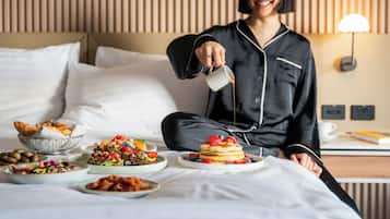 En person som häller upp en dryck i en kopp, en frukostuppsättning på ett sängbord och ett litet bord bredvid sängen med en lampa.