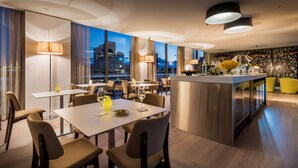 A modern dining area with tables and chairs, a central island with a bar, and large windows offering a city view. - PARKROYAL Parramatta (Parramatta)