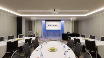 Un salón de conferencias con una pantalla grande que muestra 'PARKROYAL PARRAMATTA', mesas dispuestas con botellas de agua y manzanas, y un proyector.