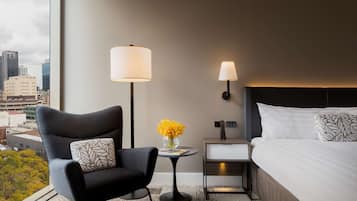 Habitación de hotel con diseño moderno, que cuenta con un amplio ventanal con vista a la ciudad, un sillón cómodo, una mesita de noche con lámpara y una cama bien hecha.