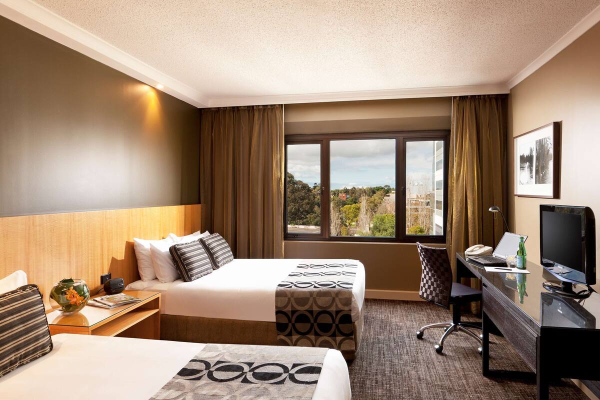 Ein Hotelzimmer mit zwei Betten, einem Schreibtisch mit Computer, einem Fernseher und einem Fenster mit Blick auf die Stadt.