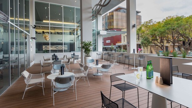 A modern outdoor seating area with tables and chairs, large windows, and a view of the cityscape. - PARKROYAL Parramatta (Parramatta)
