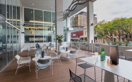 A modern outdoor seating area with tables and chairs, large windows, and a view of the cityscape.