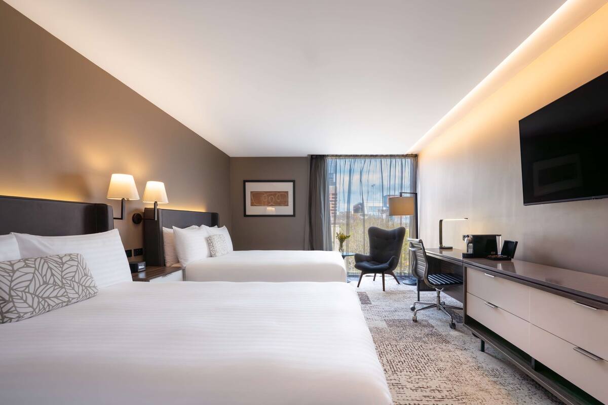 Ein Hotelzimmer mit zwei Betten, einem Schreibtisch, einem Sessel, einem Flachbildfernseher und einem Fenster mit Vorhängen.