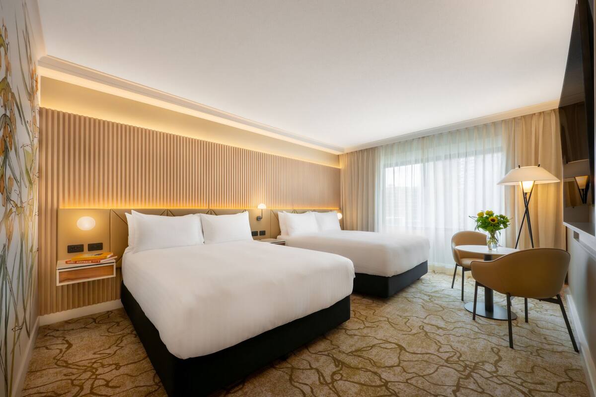 Ein Hotelzimmer mit zwei Betten, einem Schreibtisch, einem Stuhl, einer Lampe und einem fenster mit Vorhängen.