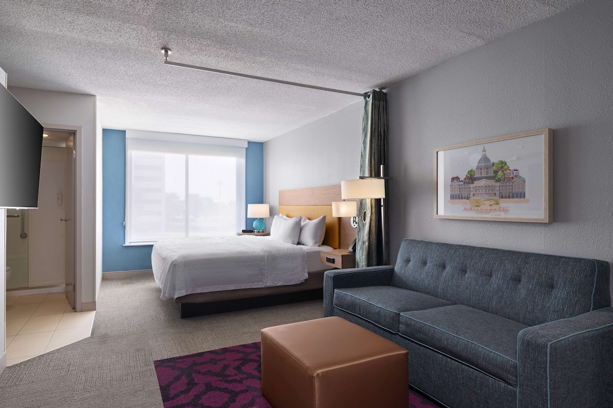 A hotel room with a bed, a grey sofa, a brown ottoman, a nightstand with a lamp, and a framed picture on the wall.