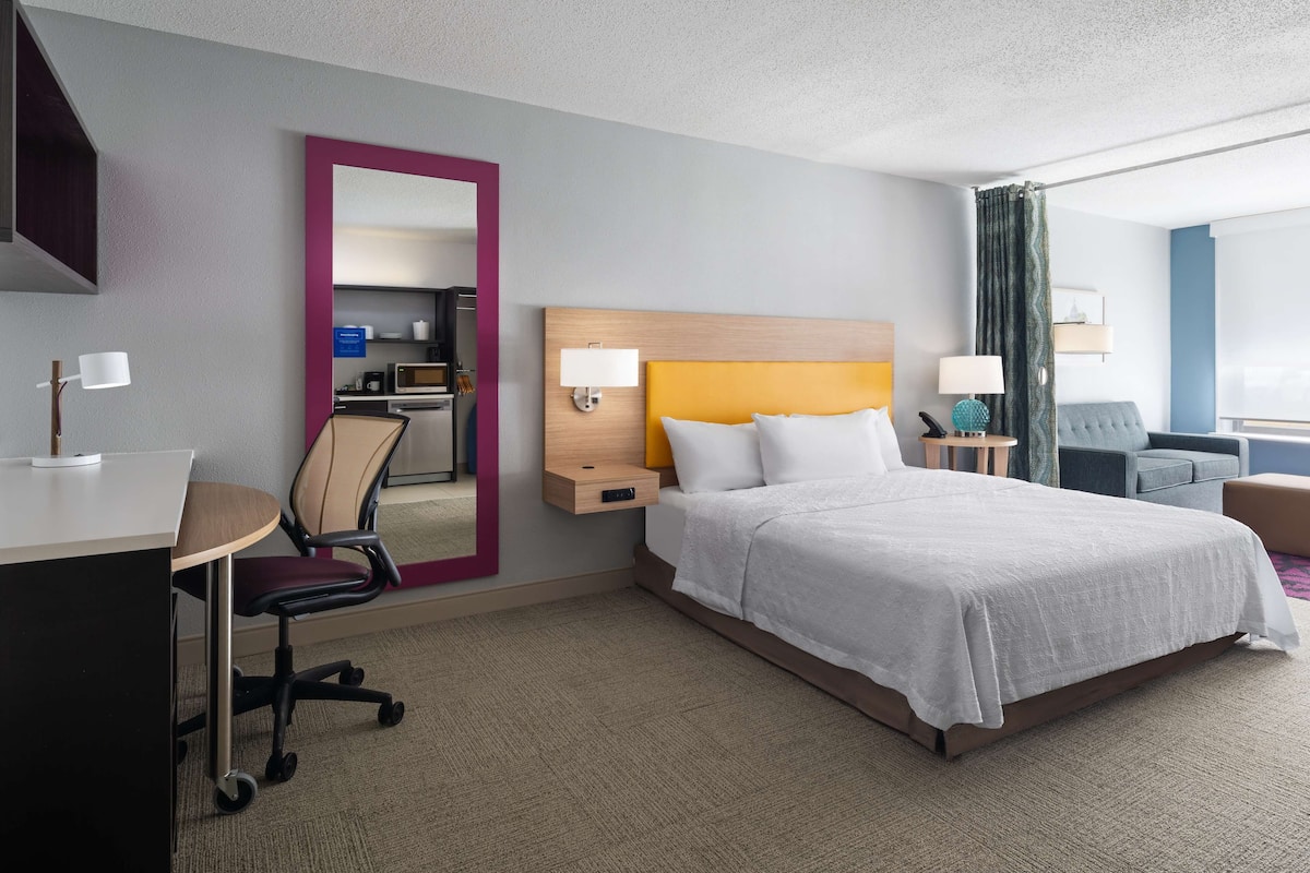 A hotel room with a bed, desk, chair, and a small kitchenette.