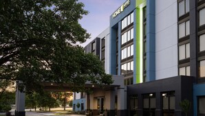 Un hotel moderno con un llamativo letrero de "Home Suites by Hilton", un acceso cubierto y un área exterior bien cuidada con árboles y un sendero.
