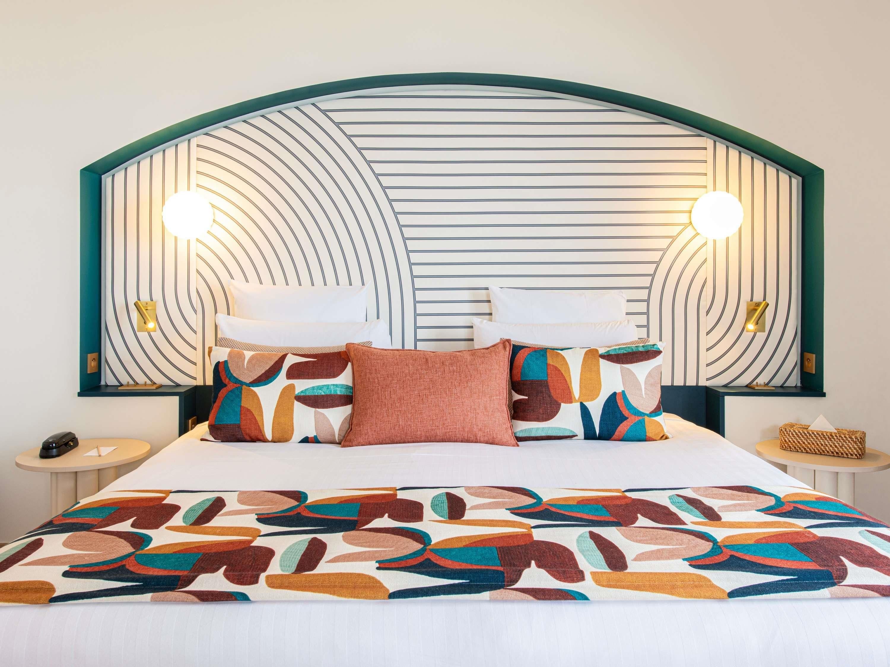 Una habitación de hotel con una cama grande, cabecera curvada, dos lámparas de noche y una manta decorativa.