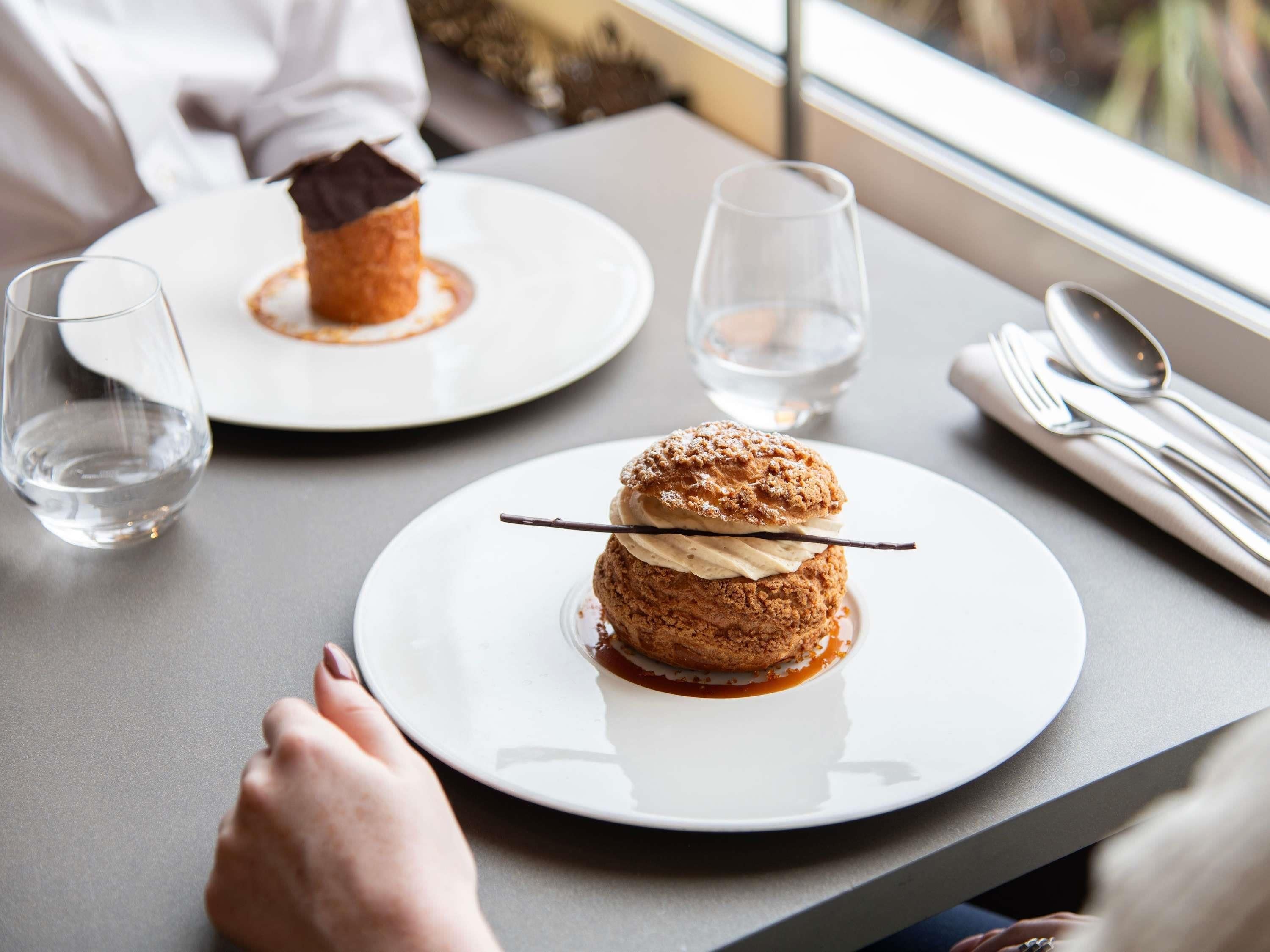 A dessert setting with two plates of dessert on a table, a glass of water, and a window view.