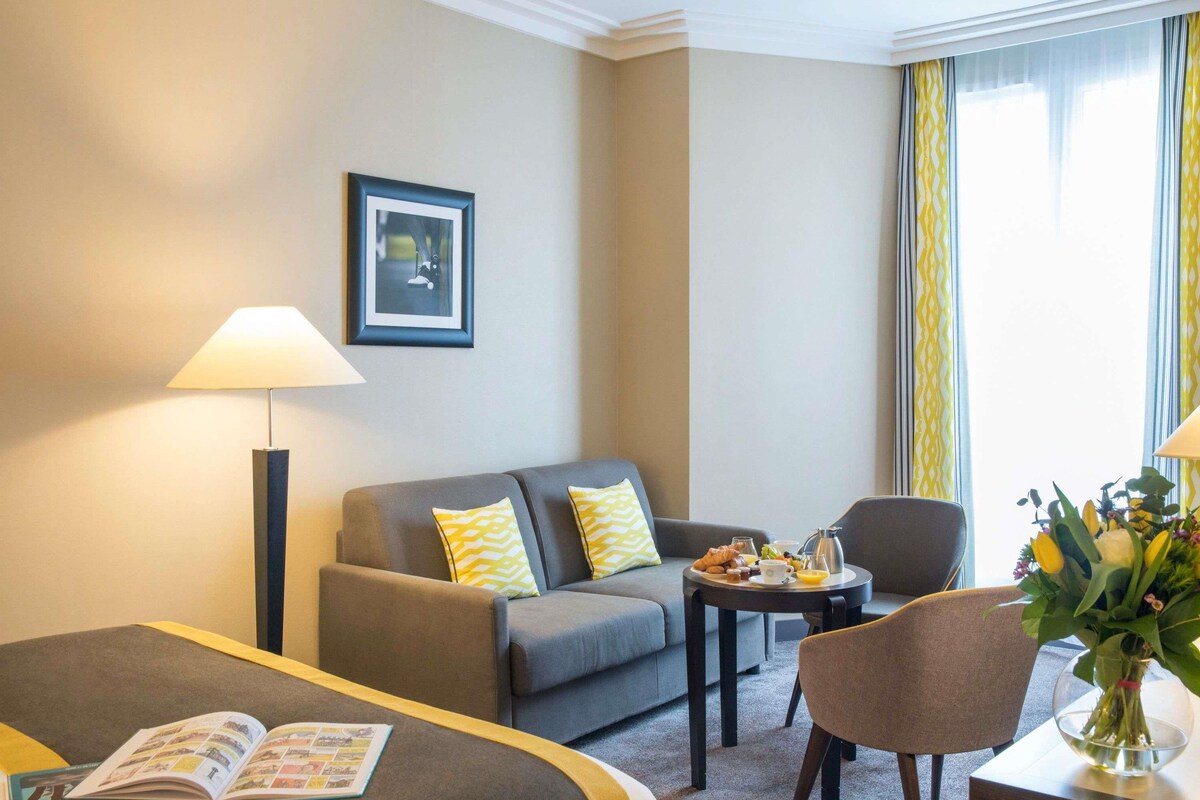 Une chambre d’hôtel avec un lit, un canapé, une petite table avec un plateau de petit-déjeuner, une lampe et un tableau au mur.