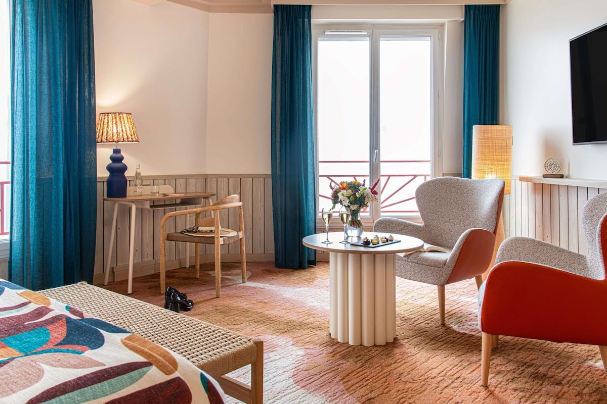 A hotel room with a balcony, a dining table, two chairs, a sofa, a TV, and a lamp.
