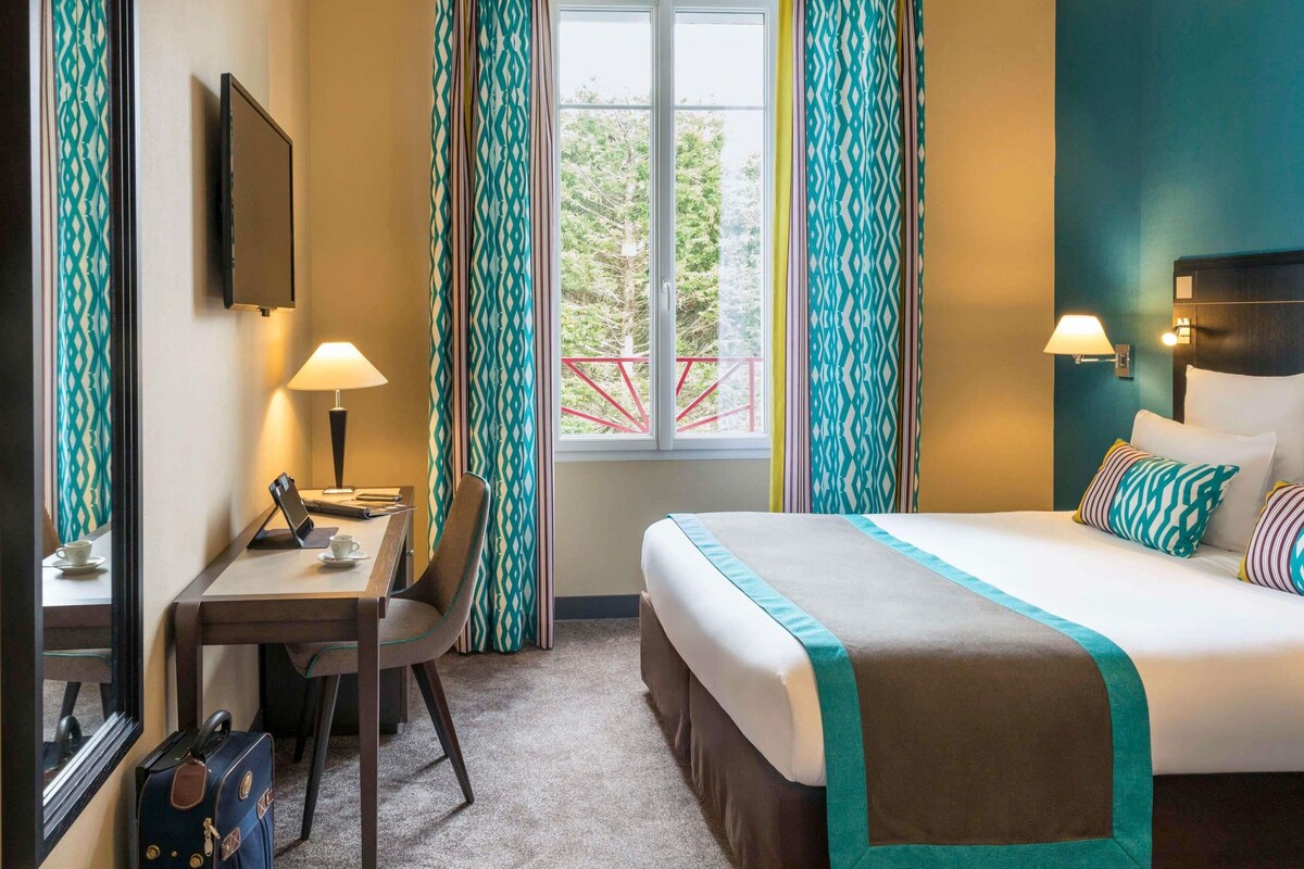 Une chambre d’hôtel avec un lit, un bureau, une chaise et une fenêtre avec des rideaux.