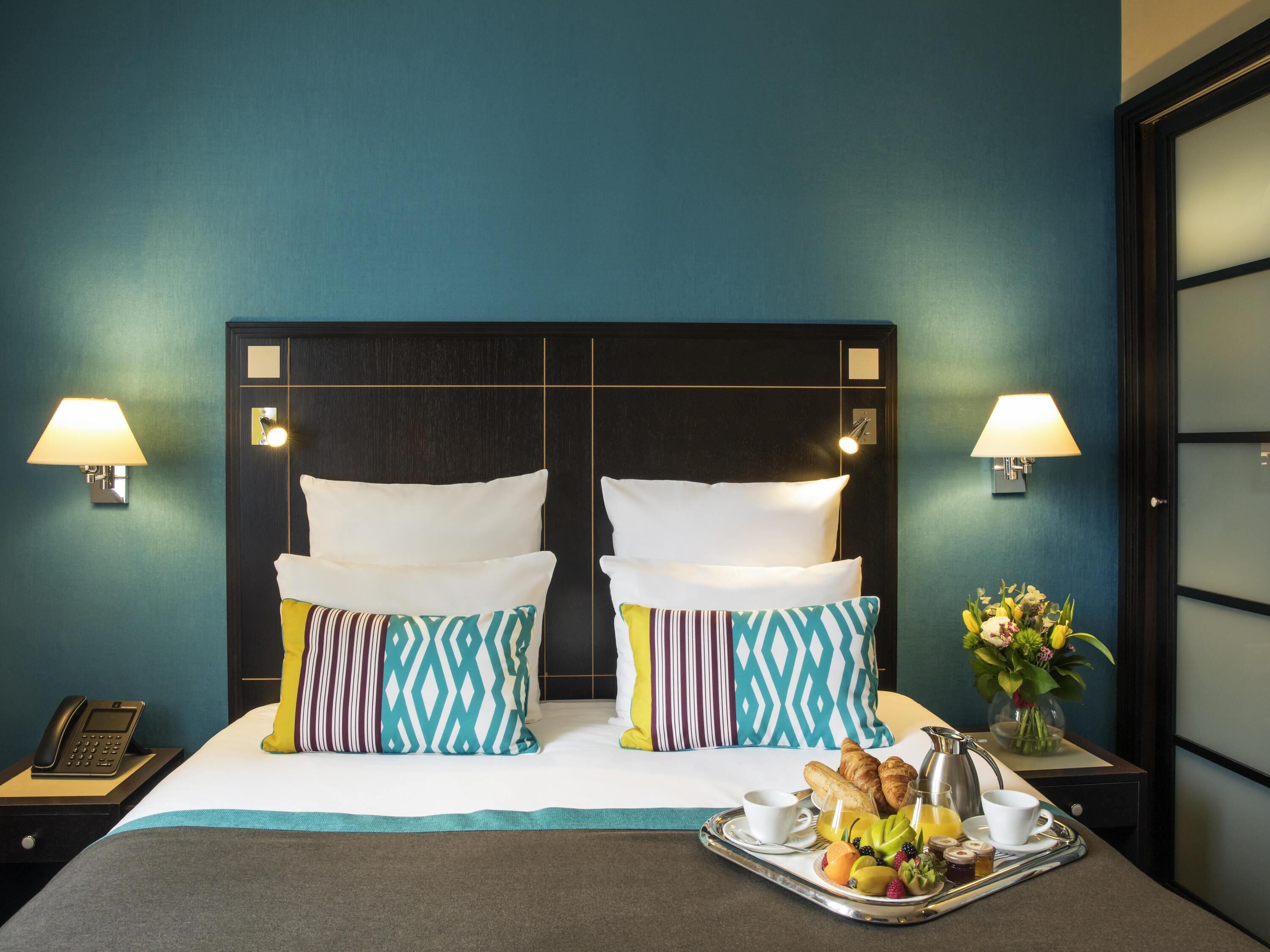 A hotel room with a large bed, two bedside lamps, a telephone, a tray with breakfast items, and a vase of flowers.
