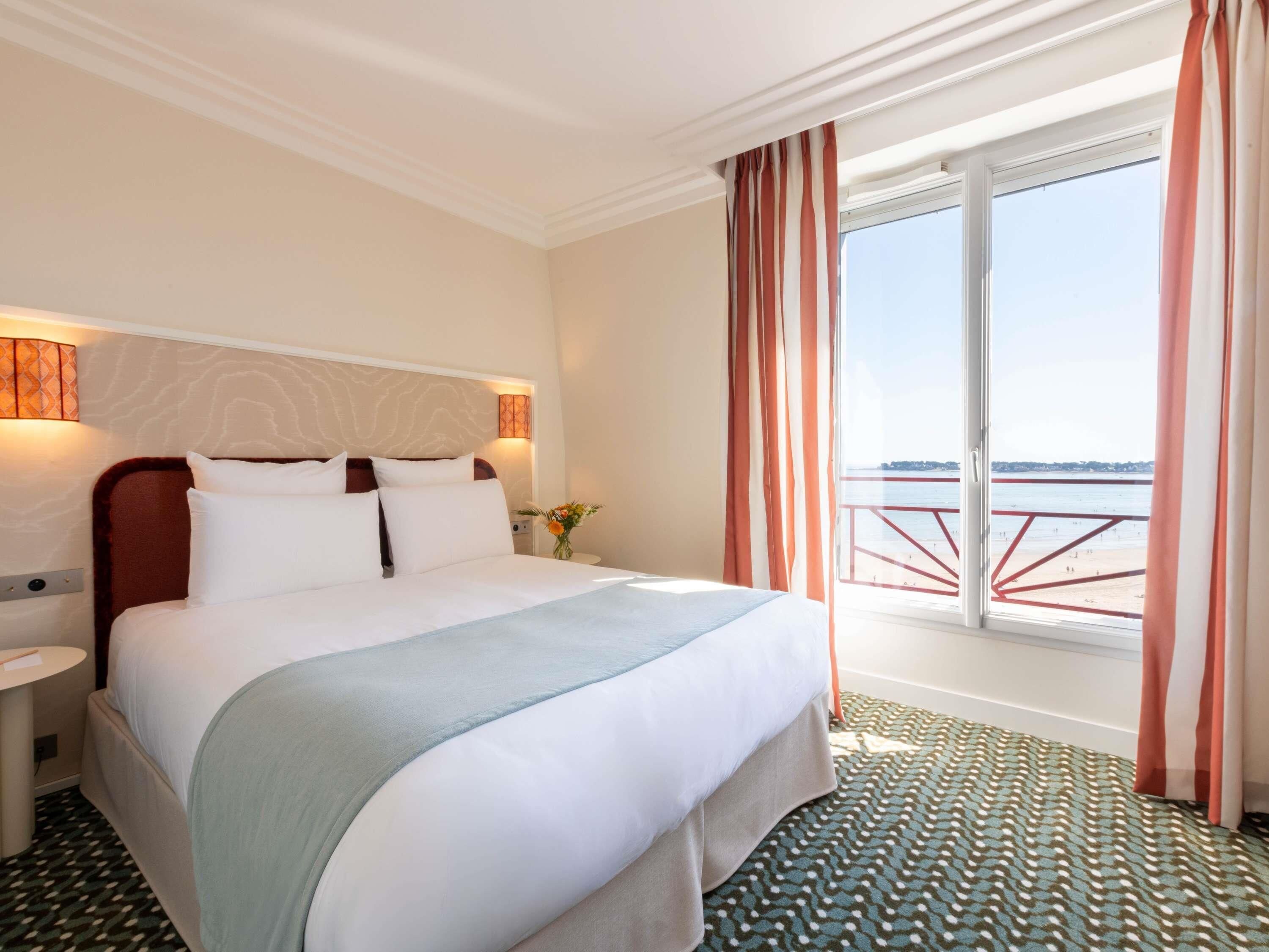A hotel room with a bed, a nightstand, a window with curtains, and a view of a pier.