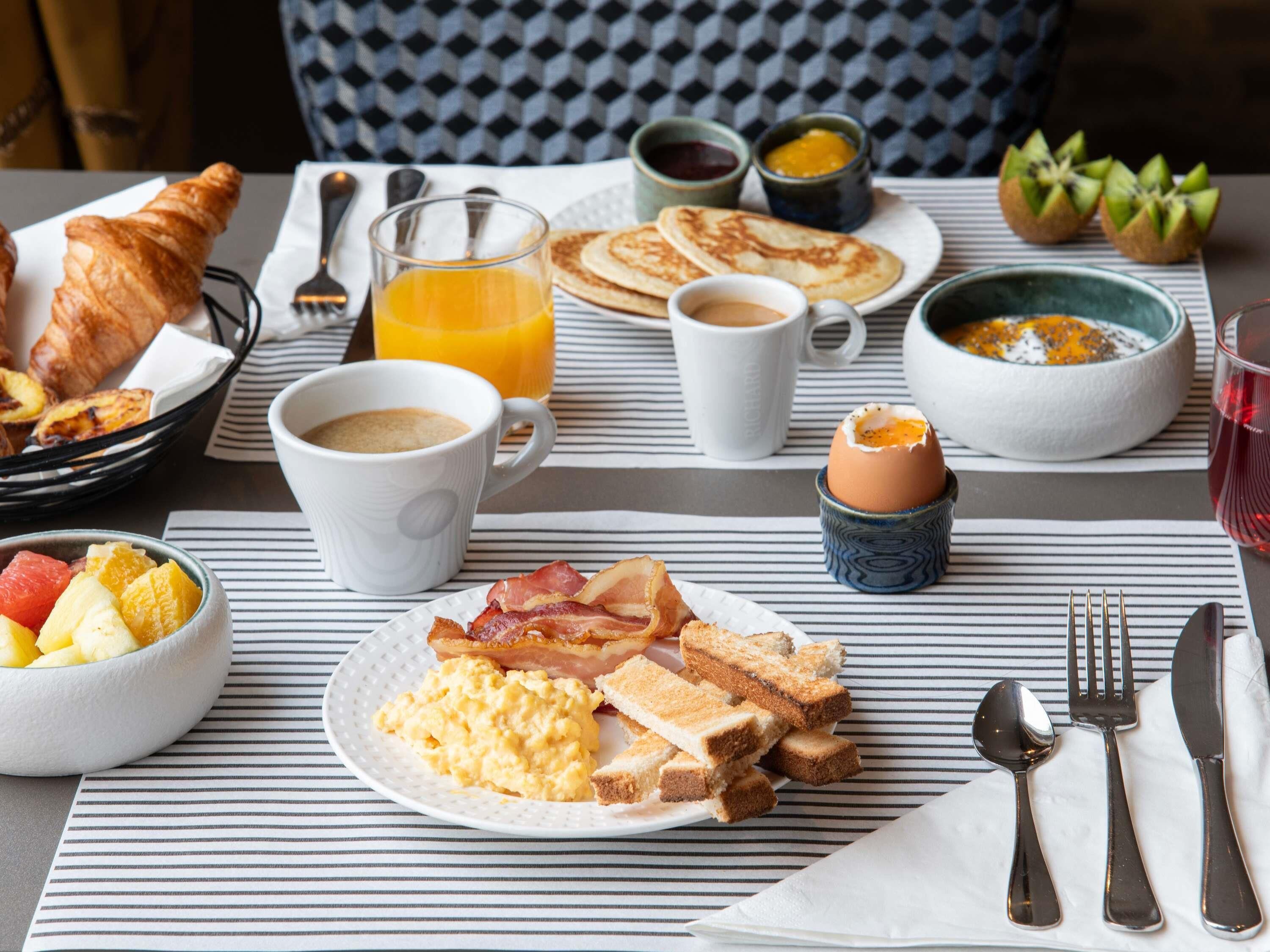 Ett frukostbord med croissanter, äggröra, bacon, rostat bröd, frukt, apelsinjuice, kaffe och olika tillbehör.