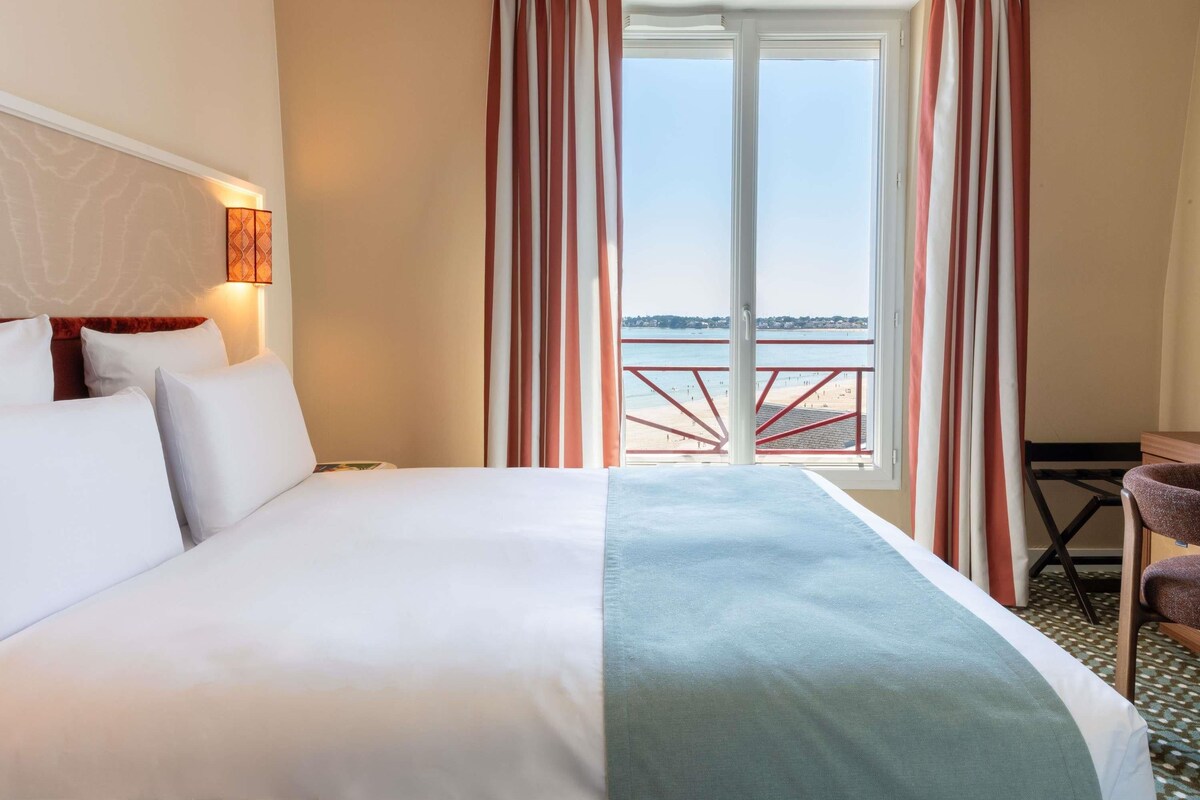 Une chambre d’hôtel avec un lit, un bureau et une vue sur la mer, grâce à de grandes fenêtres.